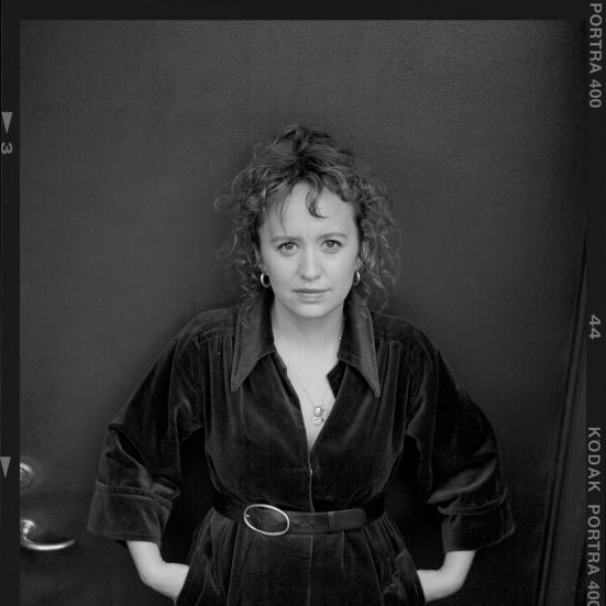 Sort-hvidt portræt af forfatteren Olga Ravn i mørk kjole med bælte, stående foran en mørk baggrund.