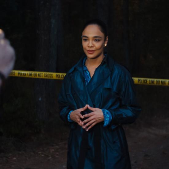 Kvinde i mørk frakke poserer foran et kamera med en politiafspærring bag hende.