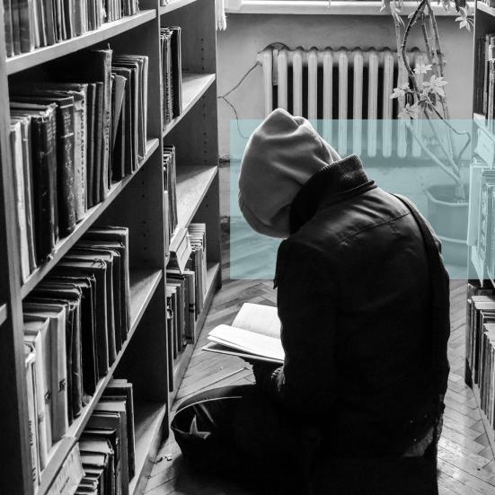 Sort-hvid foto af en person med hætte, der læser på gulvet i et bibliotek.