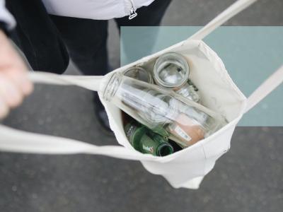 En person bærer en stofpose med tomme glasflasker og glas set oppefra.