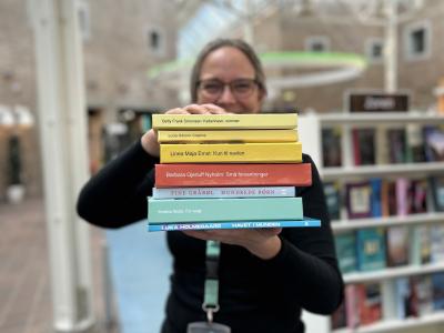 Tine står blandt fyldte bogreoler på Hovedbiblioteket. Hun holder syv af de omtalte bøger i denne artikel frem for sig i en stak med bogryggene frem. Der er fokus på bogryggene, mens både Tine og resten af baggrunden, som viser biblioteksrummet, fremstår let sløret. Man kan ane, at Tine smiler.
