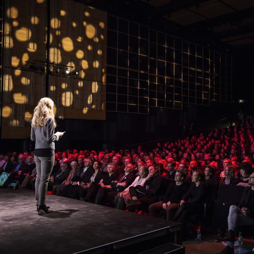 En kvinde står på scenen i Det Kongelige Biblioteks Dronningesal foran publikum.