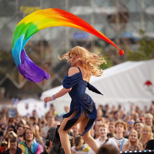 Dragperformer på scene svinger et stort regnbueflag foran en menneskemængde.