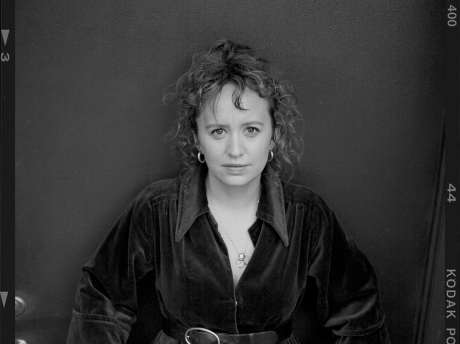 Sort-hvidt portræt af forfatteren Olga Ravn i mørk kjole med bælte, stående foran en mørk baggrund.