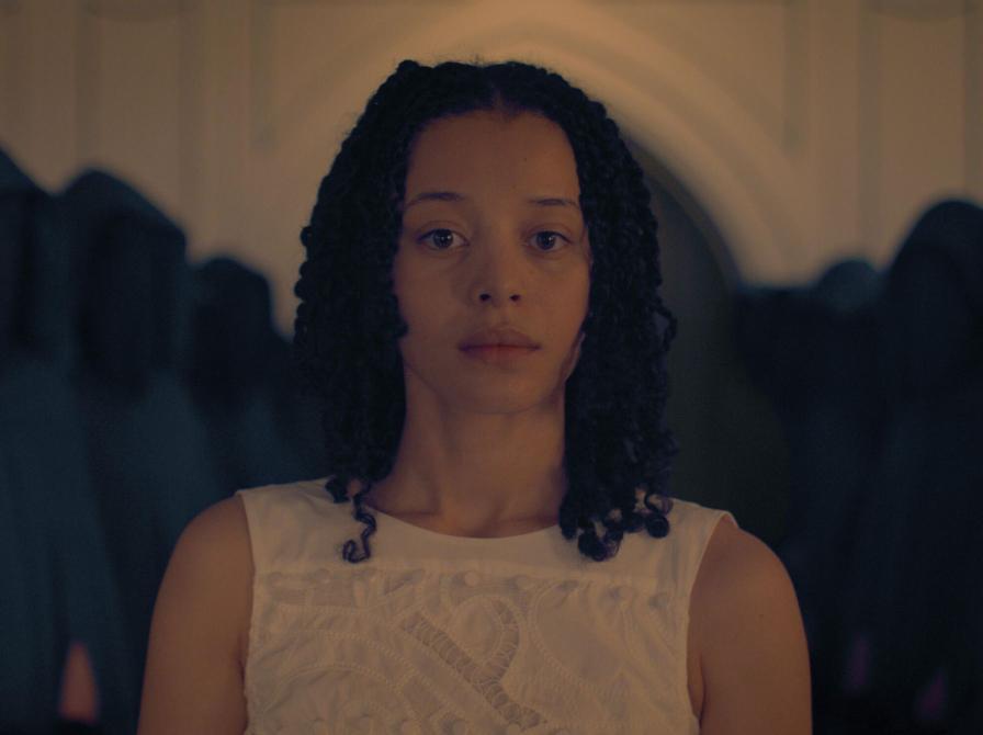 Kvinde i hvid kjole i et indendørsmiljø med mørke former i baggrunden.