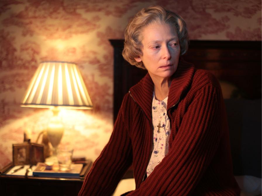 Stillbillede af Tilda Swinton fra filmen, siddende på sengekant i dæmpet belysning