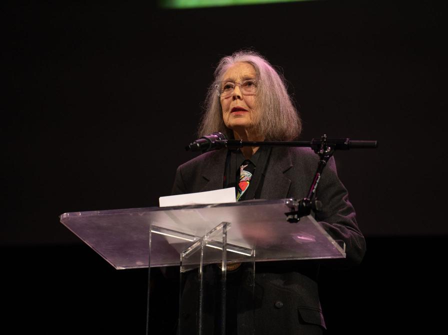 Anne Carson står på en scene ved en klar talerstol med en mikrofon foran en mørk baggrund.